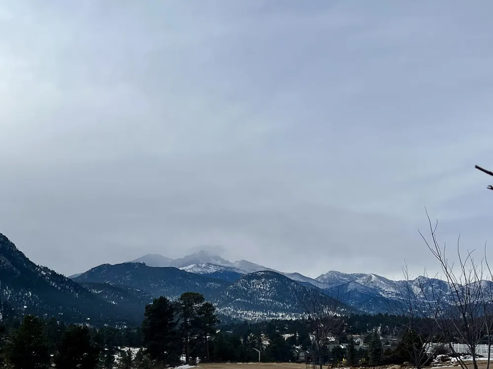 Snow-covered Longs Peak under grey cloud above the pines of Estes Park